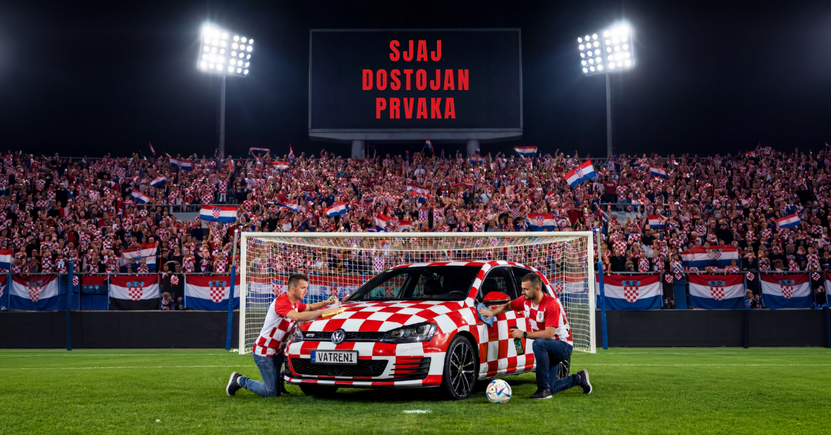 Dvojica hrvatskih navijača poliraju i čiste kockasti automobil, ispred gola na nogometnom terenu. U pozadini su hrvatski navijači.